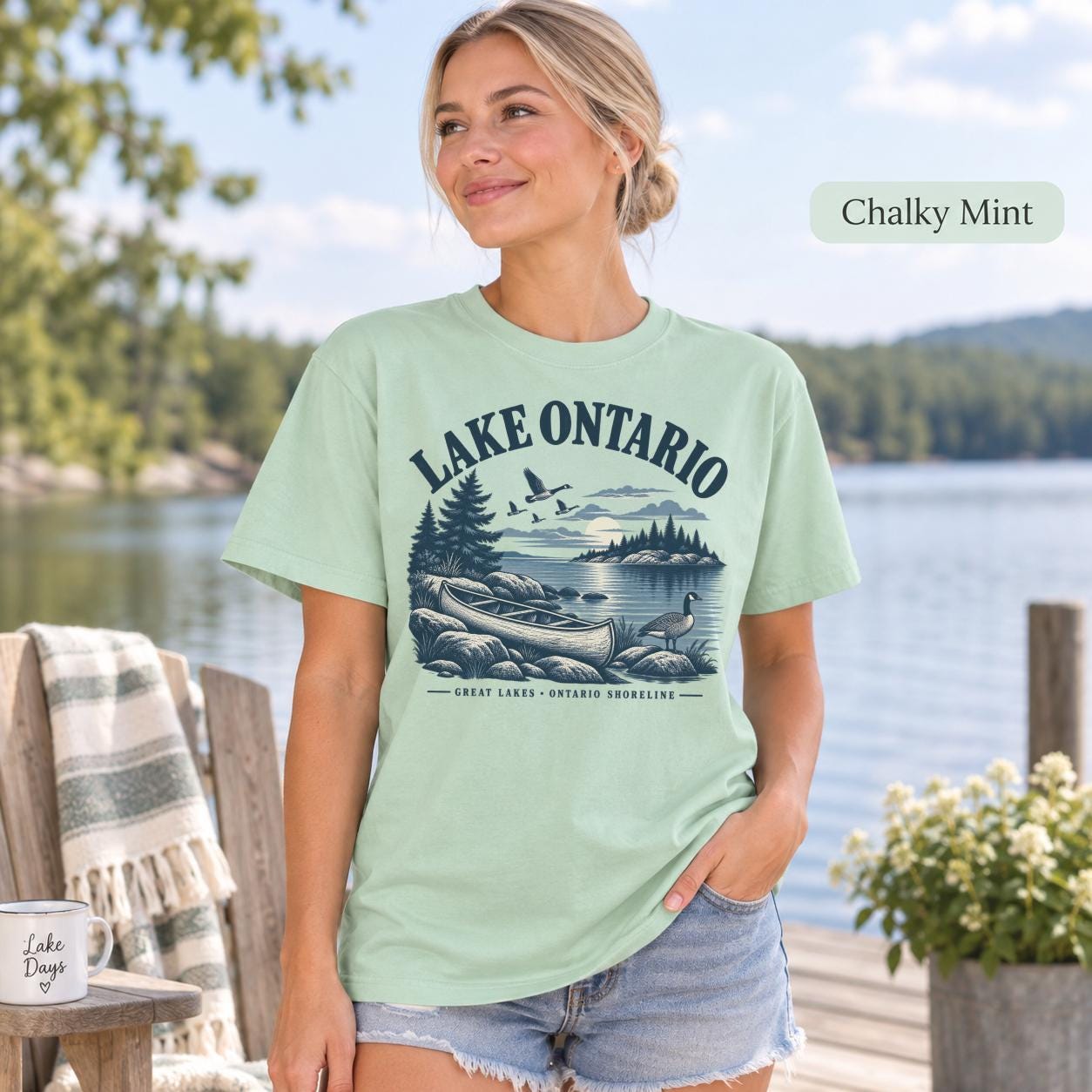 Woman wearing a Chalky Mint Comfort Colors T-shirt featuring a vintage Lake Ontario shoreline graphic with canoe, pine trees, rocky shore, Canada geese, water, and Great Lakes Ontario Shoreline text.