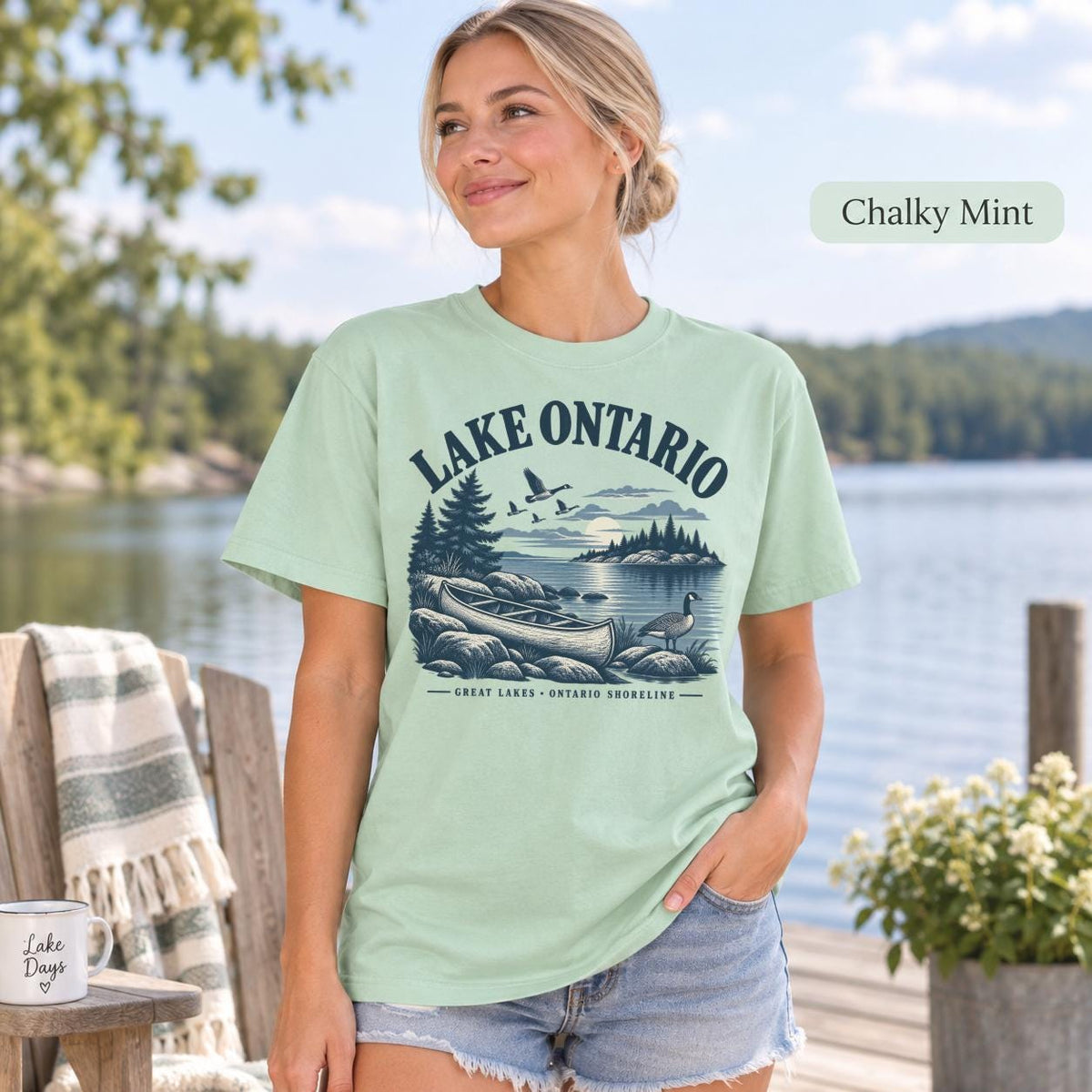 Woman wearing a Chalky Mint Comfort Colors T-shirt featuring a vintage Lake Ontario shoreline graphic with canoe, pine trees, rocky shore, Canada geese, water, and Great Lakes Ontario Shoreline text.