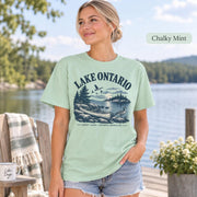 Woman wearing a Chalky Mint Comfort Colors T-shirt featuring a vintage Lake Ontario shoreline graphic with canoe, pine trees, rocky shore, Canada geese, water, and Great Lakes Ontario Shoreline text.