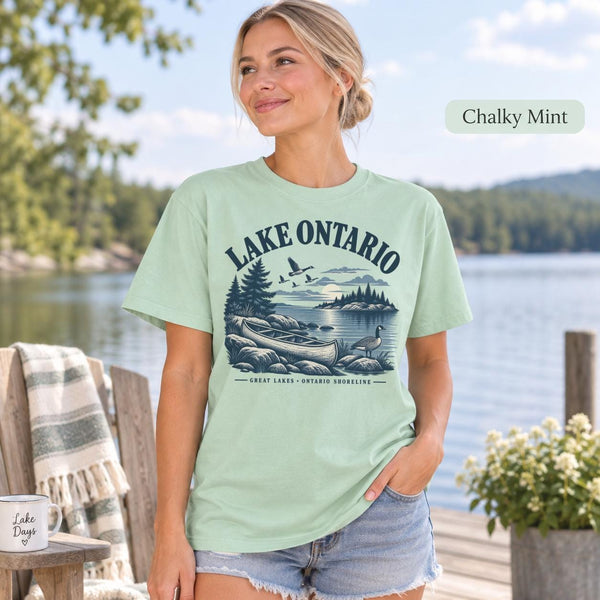 Woman wearing a Chalky Mint Comfort Colors T-shirt featuring a vintage Lake Ontario shoreline graphic with canoe, pine trees, rocky shore, Canada geese, water, and Great Lakes Ontario Shoreline text.