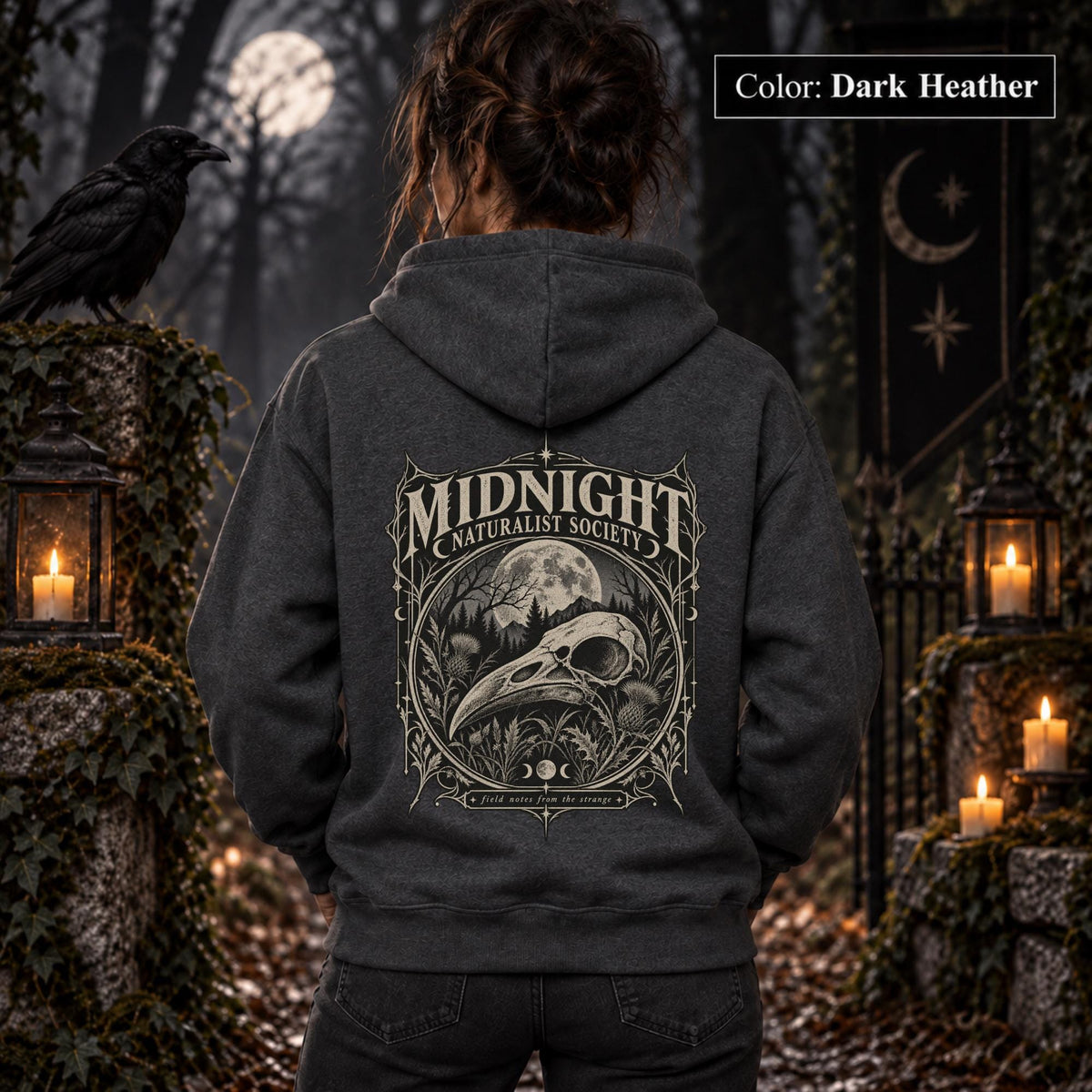 Dark Heather unisex hoodie shown from the back with a large Midnight Naturalist Society graphic featuring a bird skull, moon, mountains, forest plants, and vintage gothic botanical details.
