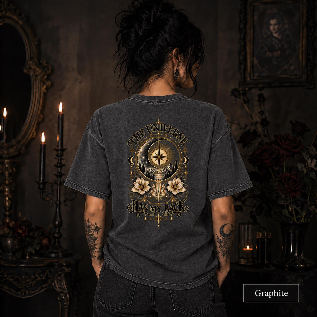 Female model wearing a Graphite Comfort Colors 1717 tee with a large gold celestial graphic reading The Universe Has My Back, featuring compass imagery, mountains, flowers, crystals, stars, and crescent moons.