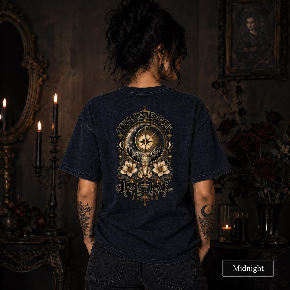 Female model wearing a Midnight Comfort Colors 1717 tee with a large gold celestial graphic reading The Universe Has My Back, featuring compass imagery, mountains, flowers, crystals, stars, and crescent moons.