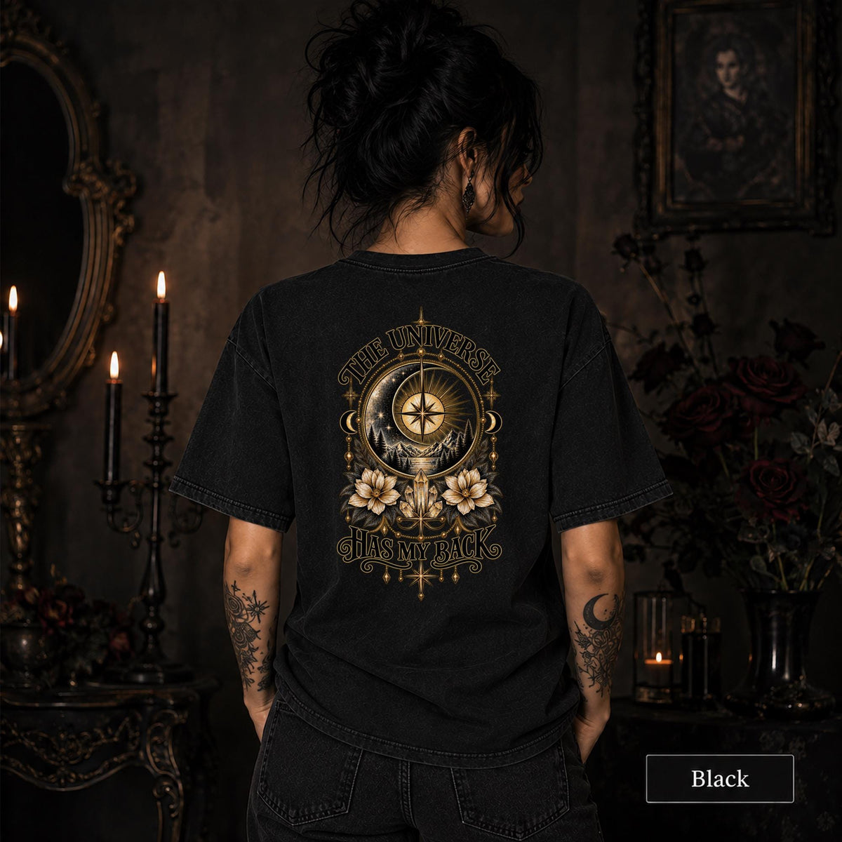Female model wearing a black Comfort Colors 1717 tee with a large gold celestial graphic reading The Universe Has My Back, featuring compass imagery, mountains, flowers, crystals, stars, and crescent moons.