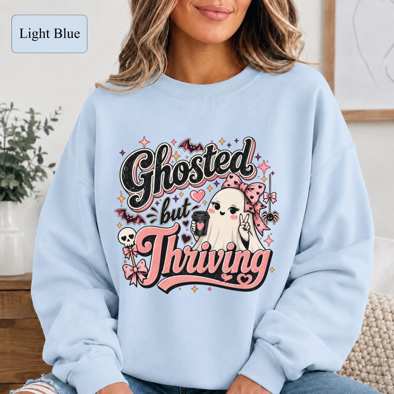 Woman wearing a light blue crewneck sweatshirt featuring a cute bow-wearing ghost holding coffee, surrounded by pink hearts, bats, stars, and the words Ghosted but Thriving.