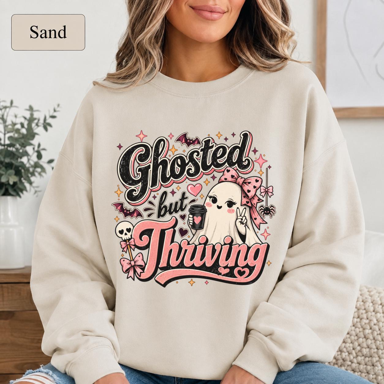 Woman wearing a sand crewneck sweatshirt featuring a cute bow-wearing ghost holding coffee, surrounded by pink hearts, bats, stars, and the words Ghosted but Thriving.