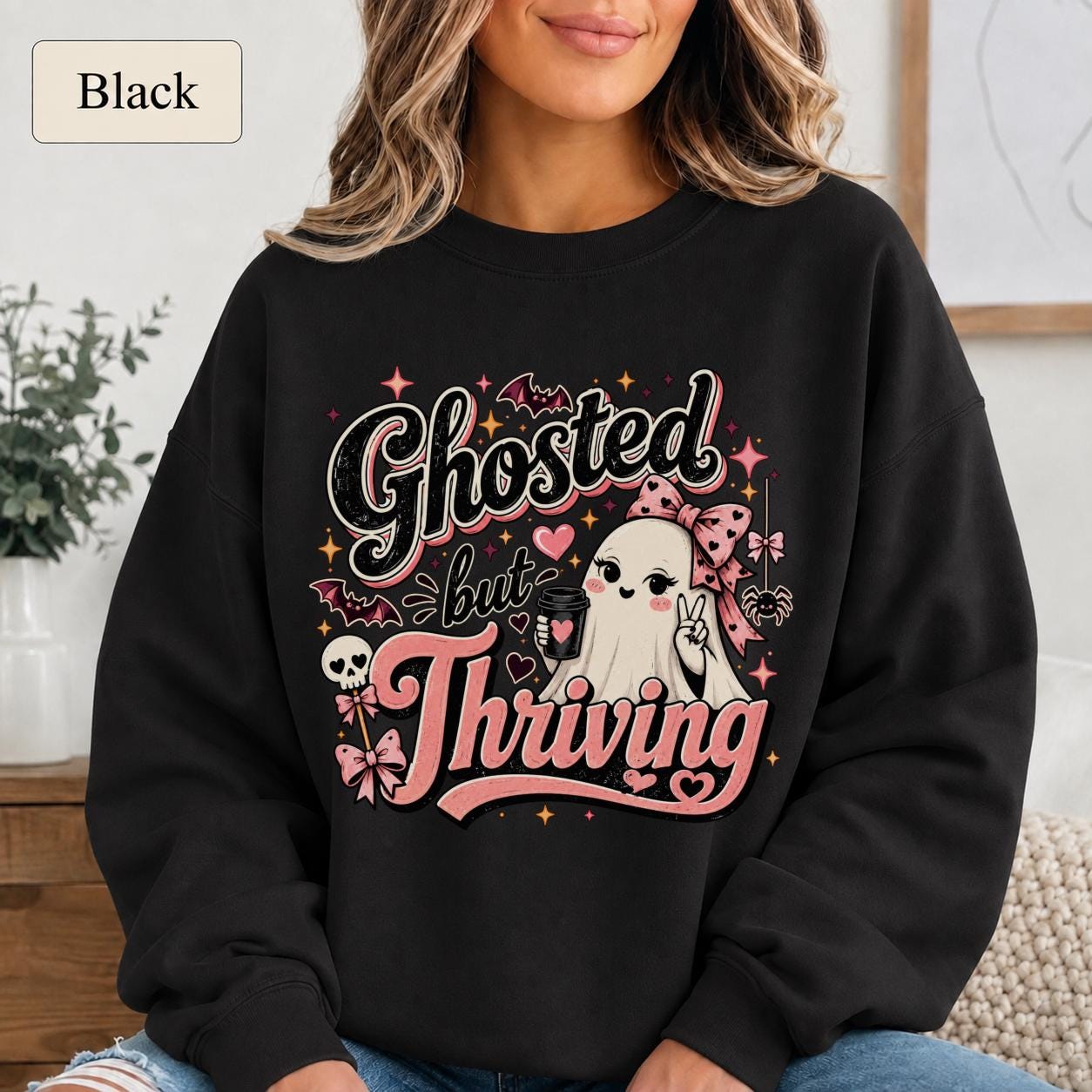 Woman wearing a black crewneck sweatshirt featuring a cute bow-wearing ghost holding coffee, surrounded by pink hearts, bats, stars, and the words Ghosted but Thriving.