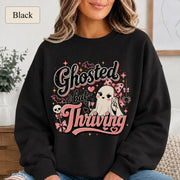 Woman wearing a black crewneck sweatshirt featuring a cute bow-wearing ghost holding coffee, surrounded by pink hearts, bats, stars, and the words Ghosted but Thriving.