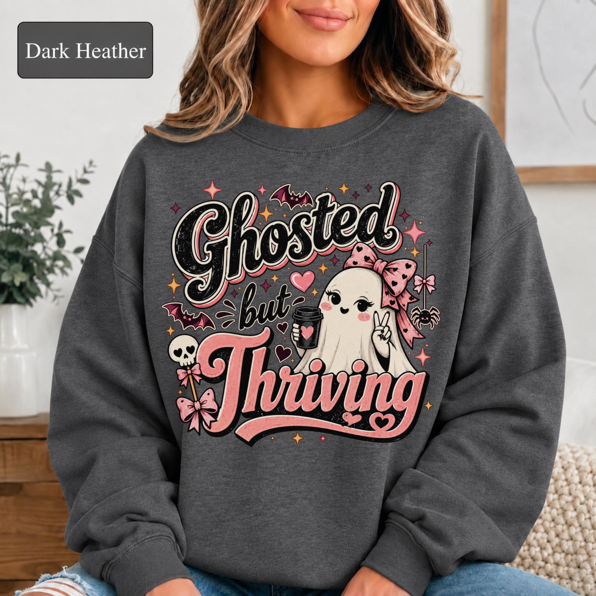 Woman wearing a dark heather crewneck sweatshirt featuring a cute bow-wearing ghost holding coffee, surrounded by pink hearts, bats, stars, and the words Ghosted but Thriving.