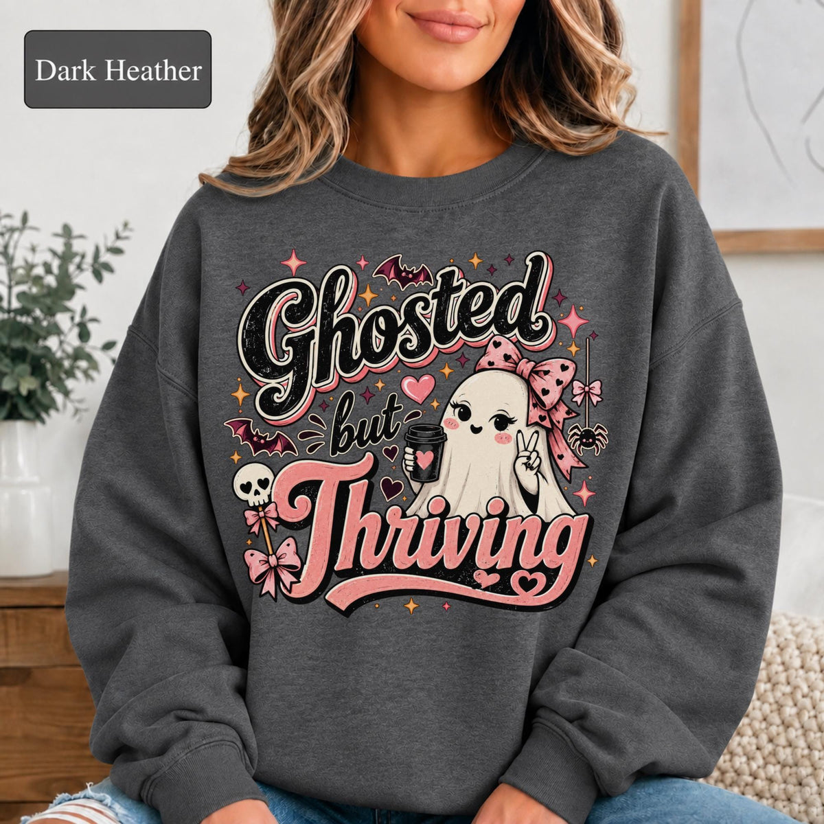Woman wearing a dark heather crewneck sweatshirt featuring a cute bow-wearing ghost holding coffee, surrounded by pink hearts, bats, stars, and the words Ghosted but Thriving.