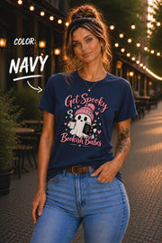 Get Spooky Bookish Babes T-shirt featuring a cute ghost in a pink beanie holding a book and coffee, with hearts and stars on a navy shirt.