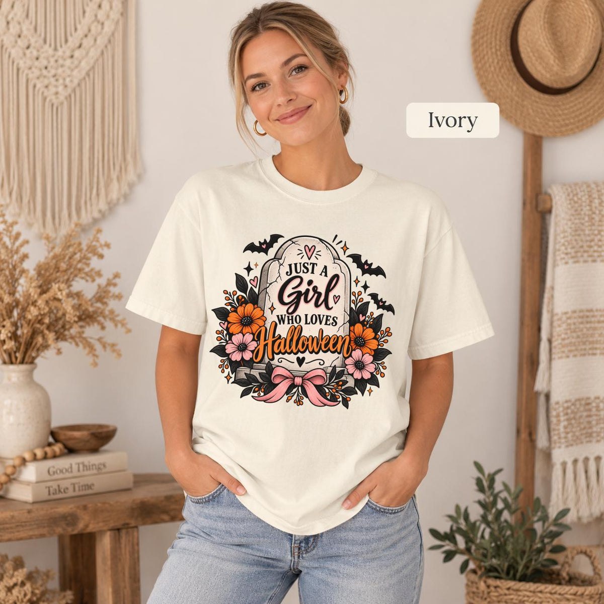 Just a Girl Who Loves Halloween T-shirt in ivory featuring a floral tombstone design with bats, orange and pink flowers, hearts, sparkles, and a pink bow.