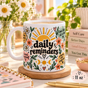 Daily Reminders ceramic mugs in 11 oz featuring colorful floral, sunshine, heart, and moon artwork with positive self-care phrases including drink water, be kind to yourself, you are enough, one step at a time, and progress over perfection.