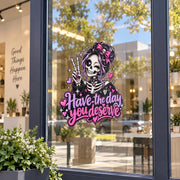 Have the Day You Deserve UV DTF decal displayed on a storefront window with gothic skeleton girl artwork in pink, purple, black, and white.