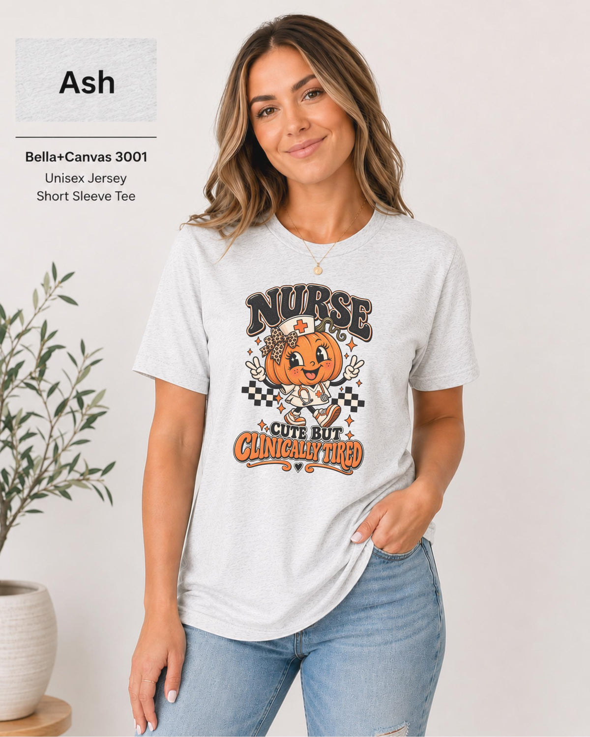 Woman wearing an Ash Bella+Canvas 3001 unisex t-shirt featuring a cute pumpkin nurse graphic and the phrase “Nurse Cute But Clinically Tired.”