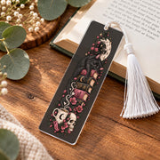 Gothic black cat bookmark with white tassel on wooden table beside an open book, decorated with pink flowers and celestial details.
