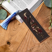 Gothic raven bookmark with blue tassel on rustic wood beside an open book, featuring moon, roses, stacked books and vintage key.
