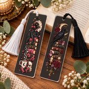 Two gothic acrylic bookmarks with tassels displayed on a rustic wooden table beside an open book, featuring a black cat design and a raven design with celestial, floral, and dark academia details.