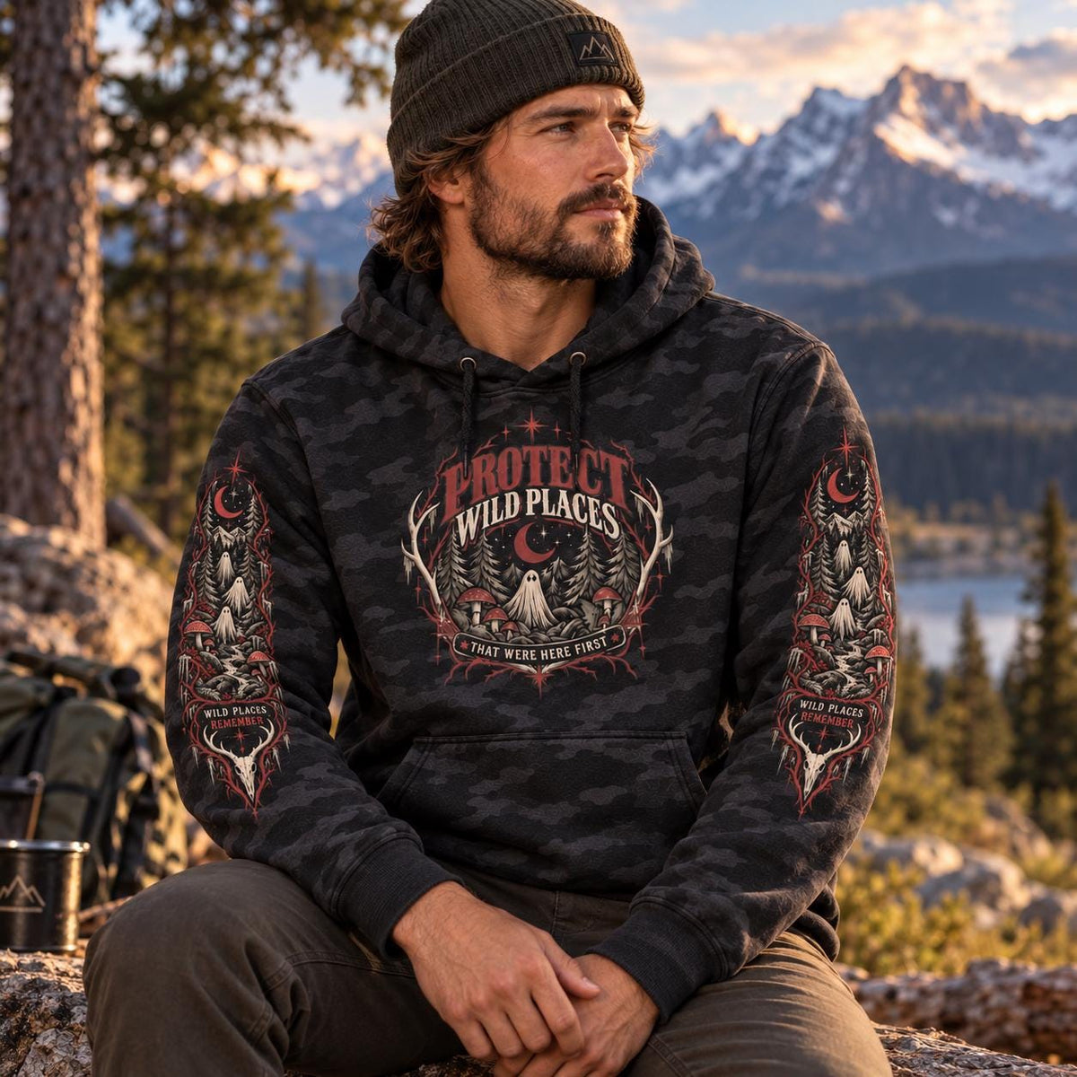 Man wearing a black camo Protect Wild Places hoodie outdoors at golden hour with mountains in the background, showing the front chest graphic and matching sleeve artwork.