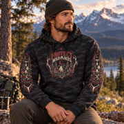 Man wearing a black camo Protect Wild Places hoodie outdoors at golden hour with mountains in the background, showing the front chest graphic and matching sleeve artwork.