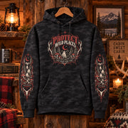 Black camo Protect Wild Places hoodie hanging in a rustic cabin setting, showing the chest design, matching sleeve graphics, kangaroo pocket, and drawstring hood.