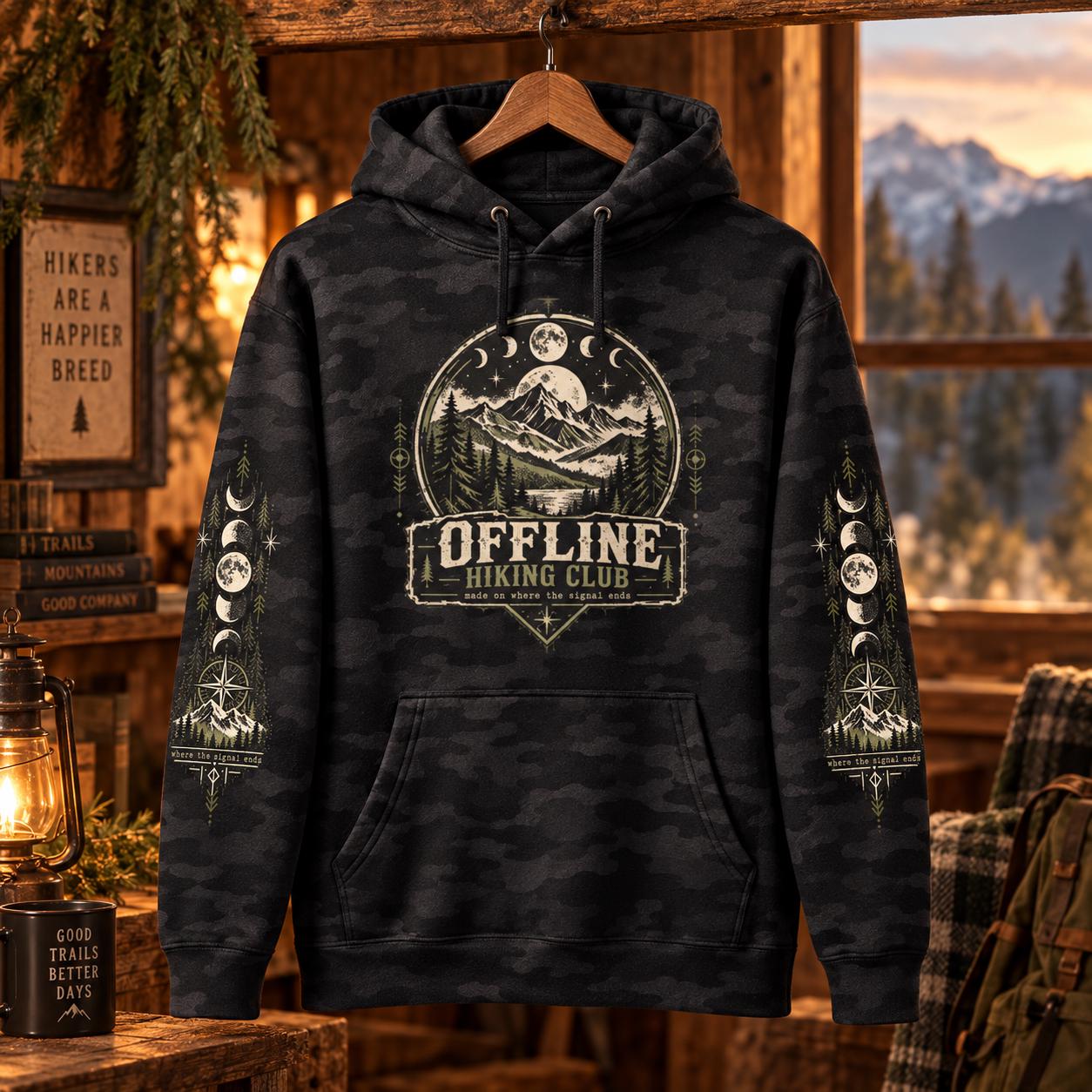 Black camo Offline Hiking Club hoodie displayed in a rustic cabin setting, featuring the front mountain graphic and coordinated sleeve artwork.