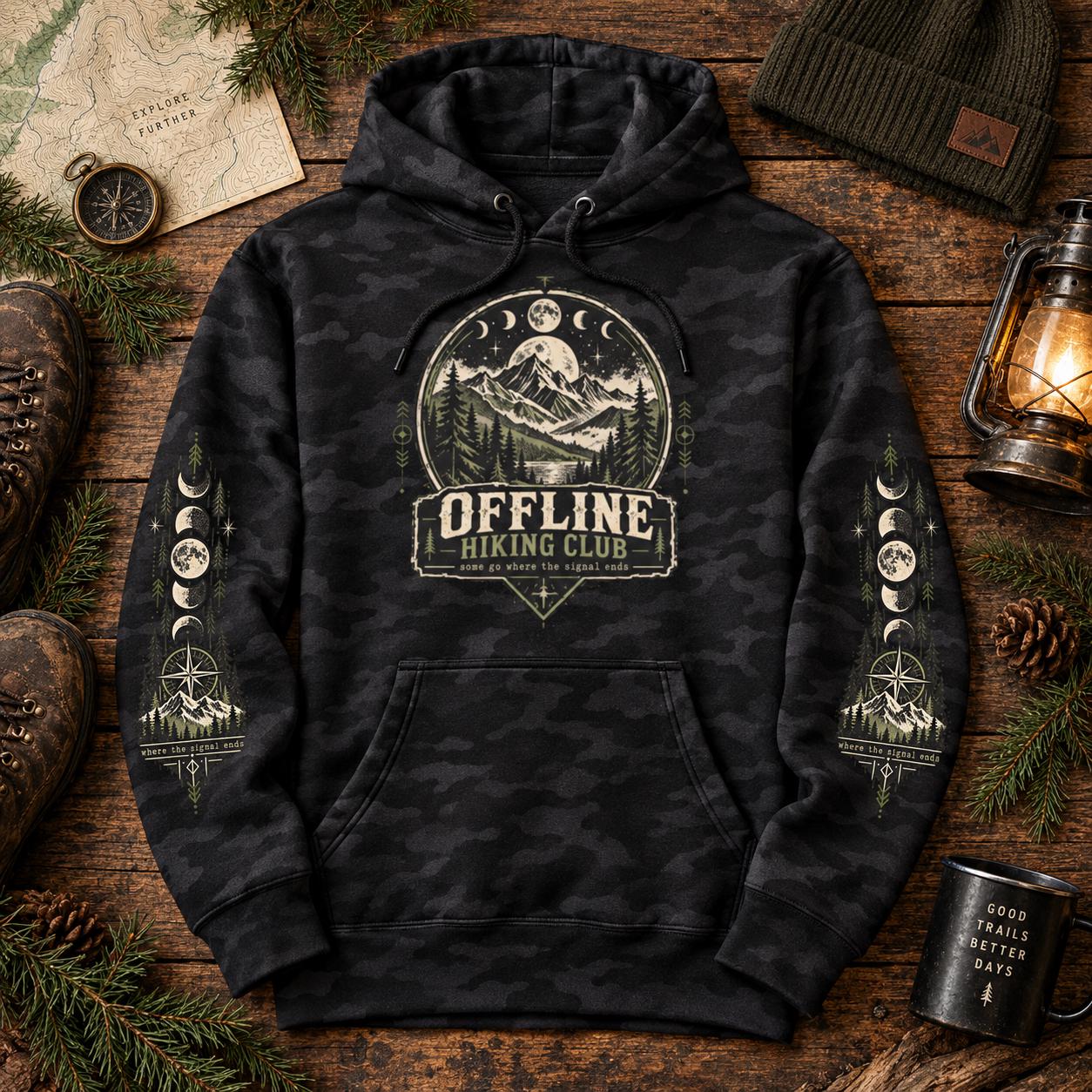 Flat lay of a black camo Offline Hiking Club hoodie with mountain, moon, compass, and pine tree design, styled with hiking gear and outdoor accessories.