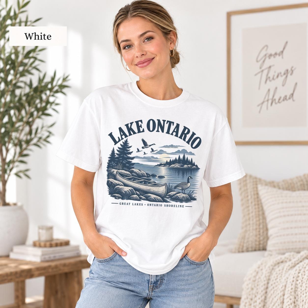 Woman wearing a white Comfort Colors T-shirt featuring a vintage Lake Ontario shoreline graphic with canoe, pine trees, rocky shore, Canada geese, water, and Great Lakes Ontario Shoreline text.