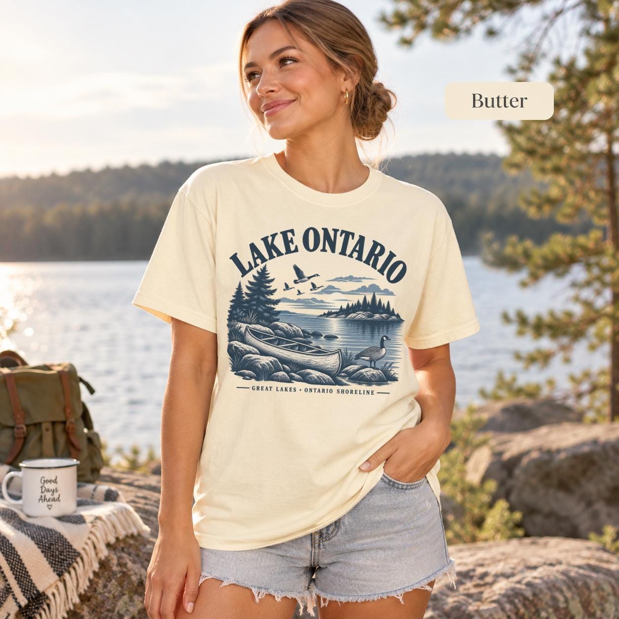 Woman wearing a Butter Comfort Colors T-shirt featuring a vintage Lake Ontario shoreline graphic with canoe, pine trees, rocky shore, Canada geese, water, and Great Lakes Ontario Shoreline text.