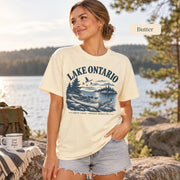 Woman wearing a Butter Comfort Colors T-shirt featuring a vintage Lake Ontario shoreline graphic with canoe, pine trees, rocky shore, Canada geese, water, and Great Lakes Ontario Shoreline text.