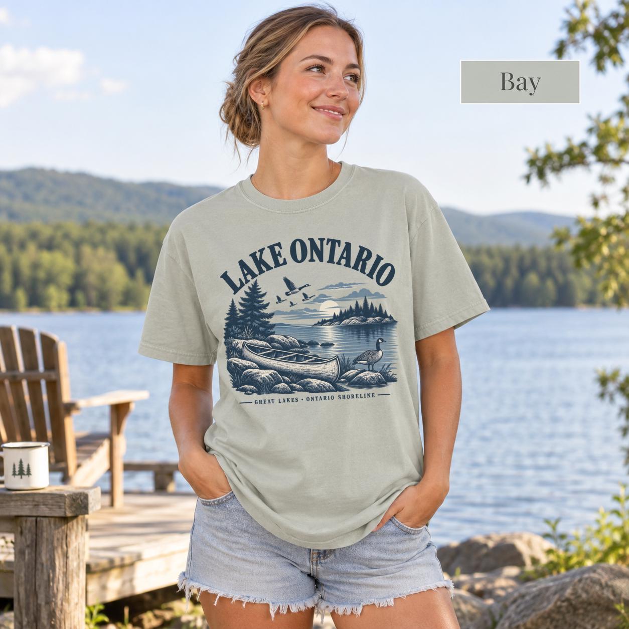 Woman wearing a Bay Comfort Colors T-shirt featuring a vintage Lake Ontario shoreline graphic with canoe, pine trees, rocky shore, Canada geese, water, and Great Lakes Ontario Shoreline text.