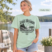 Chalky Mint Comfort Colors Lake Ontario T-shirt featuring a black vintage shoreline design with pine trees, rocky waterfront, Canada goose, Indigenous-inspired geometric details, and the phrase You Can’t Rename What Isn’t Yours.