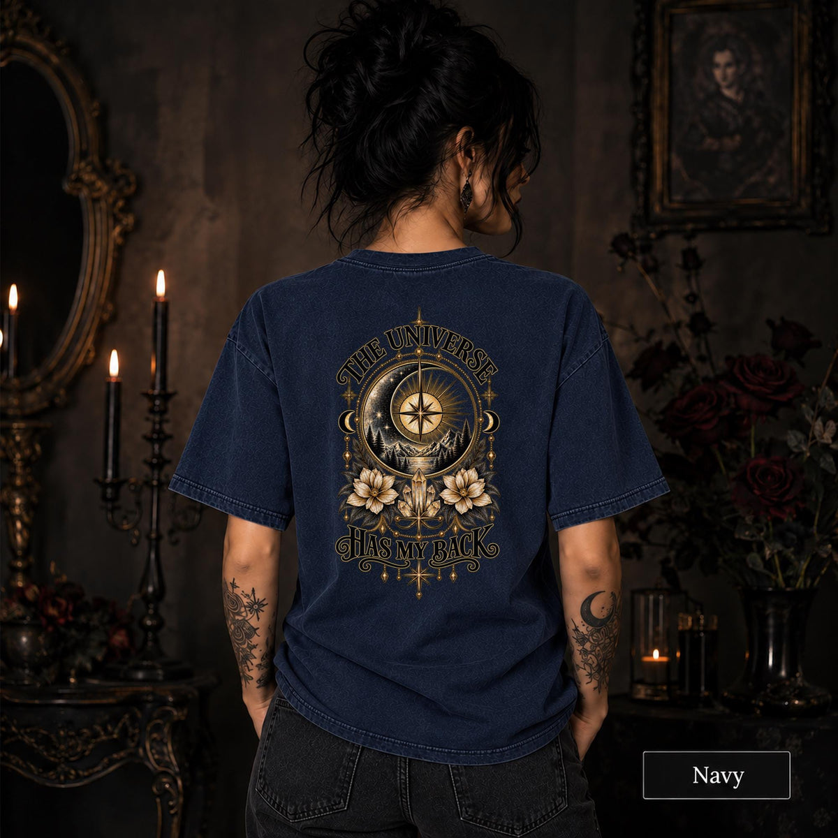 Female model wearing a navy Comfort Colors 1717 tee with a large gold celestial graphic reading The Universe Has My Back, featuring compass imagery, mountains, flowers, crystals, stars, and crescent moons.
