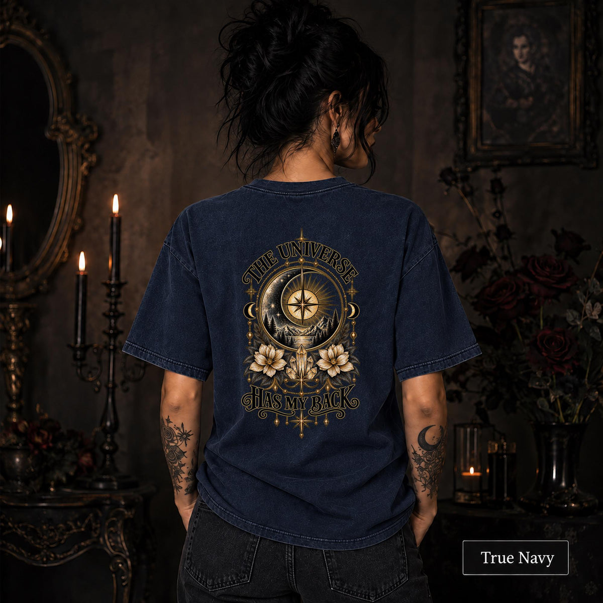 Female model wearing a True Navy Comfort Colors 1717 tee with a large gold celestial graphic reading The Universe Has My Back, featuring compass imagery, mountains, flowers, crystals, stars, and crescent moons.