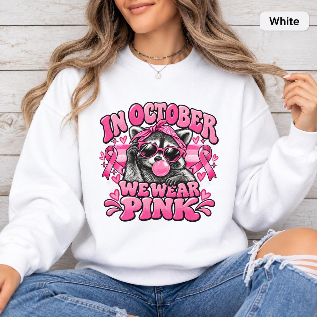 White unisex sweatshirt displaying the pink breast cancer awareness raccoon graphic with bold retro lettering and ribbon details.