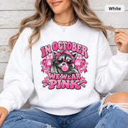 White unisex sweatshirt displaying the pink breast cancer awareness raccoon graphic with bold retro lettering and ribbon details.