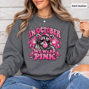 Dark Heather crewneck displaying the breast cancer support graphic with pink ribbons, hearts, and playful raccoon illustration.