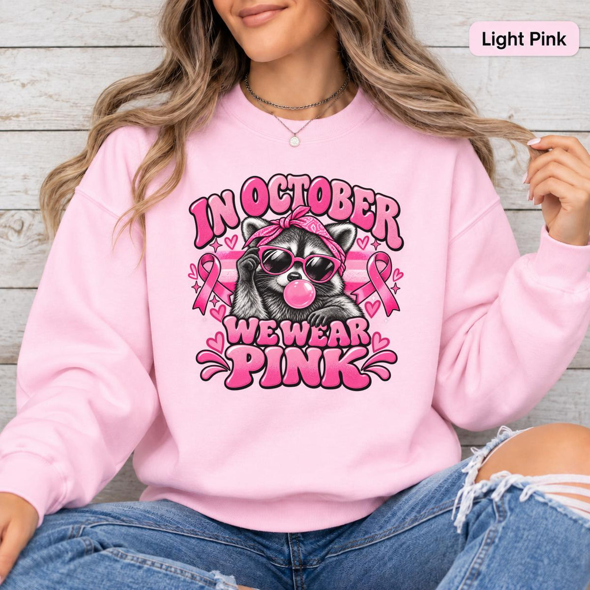 Ash crewneck sweatshirt featuring a retro raccoon in sunglasses with pink ribbons, hearts, bubblegum, and the phrase “In October We Wear Pink.