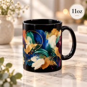 Black ceramic 11oz mug featuring colorful abstract brushstrokes in teal, blue, cream, gold, burgundy, and turquoise on a bright neutral tabletop.