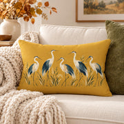 Mustard yellow lumbar pillow with five painted herons and marsh grass design styled on a cream sofa with chunky knit blanket and warm neutral autumn decor.