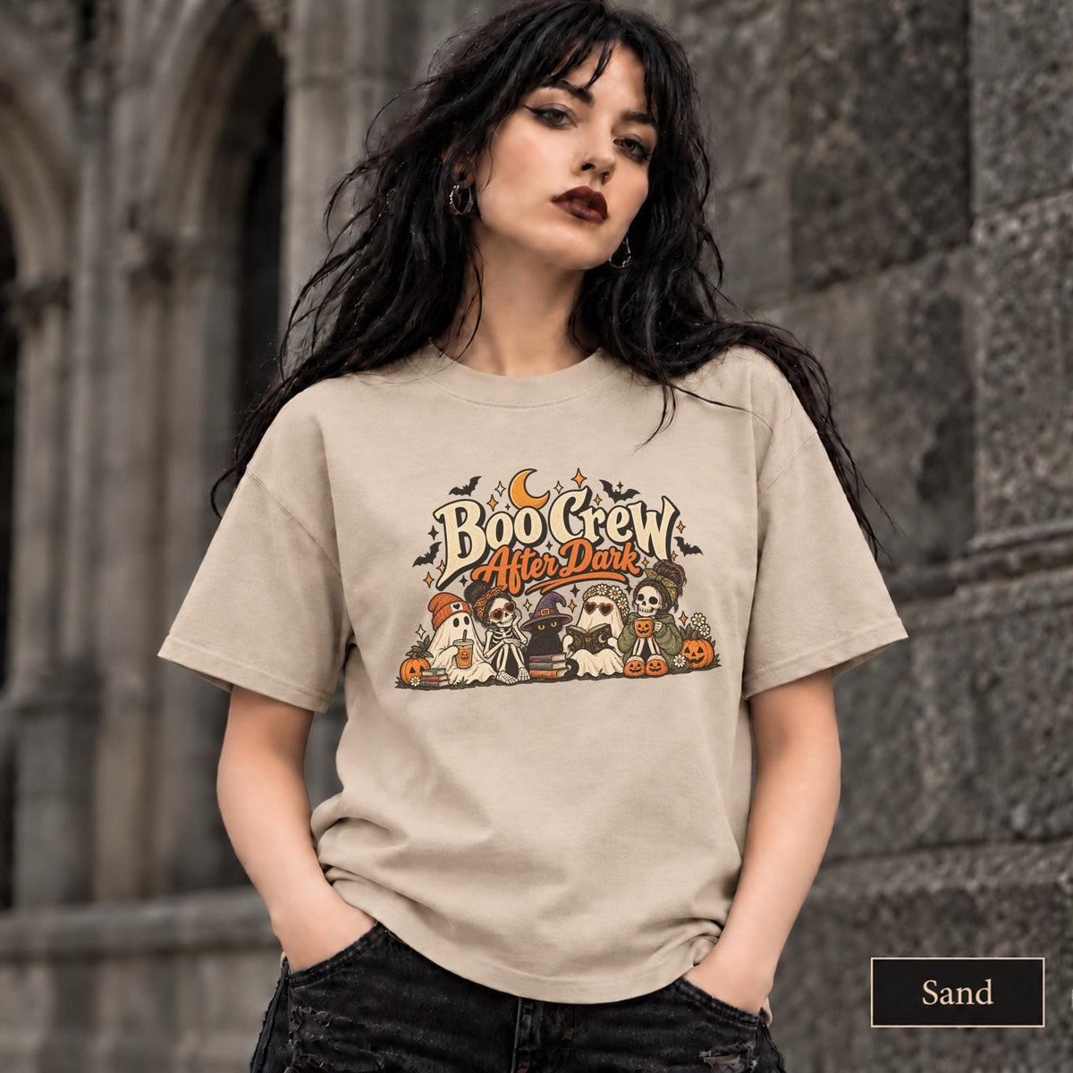 Sand unisex T-shirt featuring a “Boo Crew After Dark” Halloween design with ghosts, skeletons, pumpkins, books, bats, drinks, and a crescent moon in retro orange and cream tones.