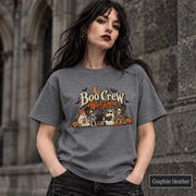 Graphite Heather unisex T-shirt featuring a “Boo Crew After Dark” Halloween design with ghosts, skeletons, pumpkins, books, bats, drinks, and a crescent moon in retro orange and cream tones.