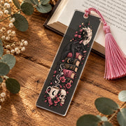 Gothic black cat bookmark with pink tassel on wooden table beside an open book, decorated with pink flowers and celestial details.