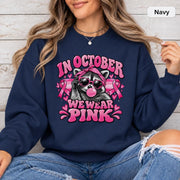 Navy crewneck mockup featuring the “In October We Wear Pink” raccoon design with pink awareness ribbons and hearts.