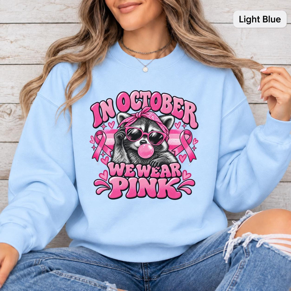 Light blue sweatshirt showcasing a playful raccoon wearing sunglasses with breast cancer awareness graphics and October pink message.