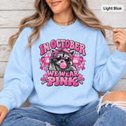 Light blue sweatshirt showcasing a playful raccoon wearing sunglasses with breast cancer awareness graphics and October pink message.