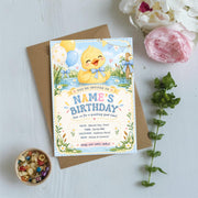 Printable duckling birthday invitation resting on a neutral tabletop with pink flowers and greenery, featuring a yellow baby duck and blue birthday details.