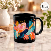 Black ceramic mug featuring bold abstract brushstrokes in coral, turquoise, orange, cream, pink, and teal on a bright neutral tabletop.