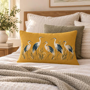 Mustard yellow heron lumbar pillow styled on a cozy neutral bed with cream bedding, textured pillows, knit throw, and soft natural light.
