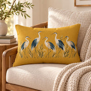 Mustard heron lumbar pillow displayed on a cream upholstered chair with warm wood accents, neutral knit blanket, greenery, and cozy home decor.